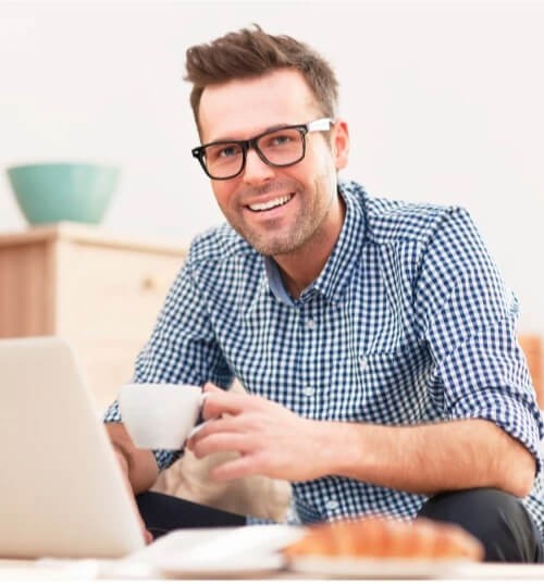 A man enjoying a coffee as he works on his laptop, while also glancing at the camera.